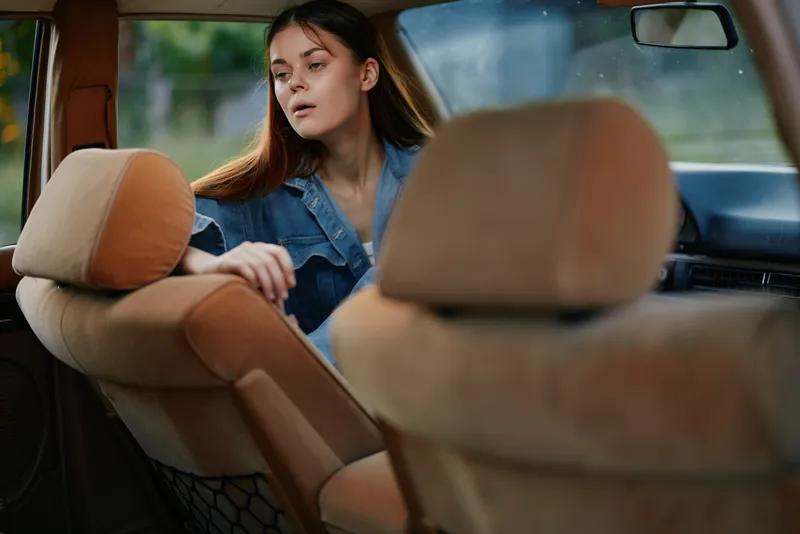A contemplative young woman in a denim jacket sitting in a car's back seat, gazing out the window, evoking feelings of introspection and serenity.