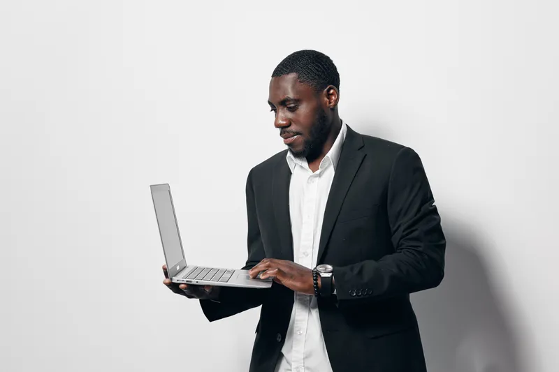 Black man with dark skin wearing black suit and white shirt using laptop for work or study on white background. Business professional, technology and communication concept with focused expression.