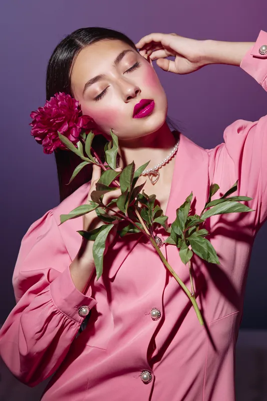 Young woman in a pink blouse holds a flower close to her face with closed eyes. Fashion makeup features bold lipstick and bright blush. Studio shot on a purple background.