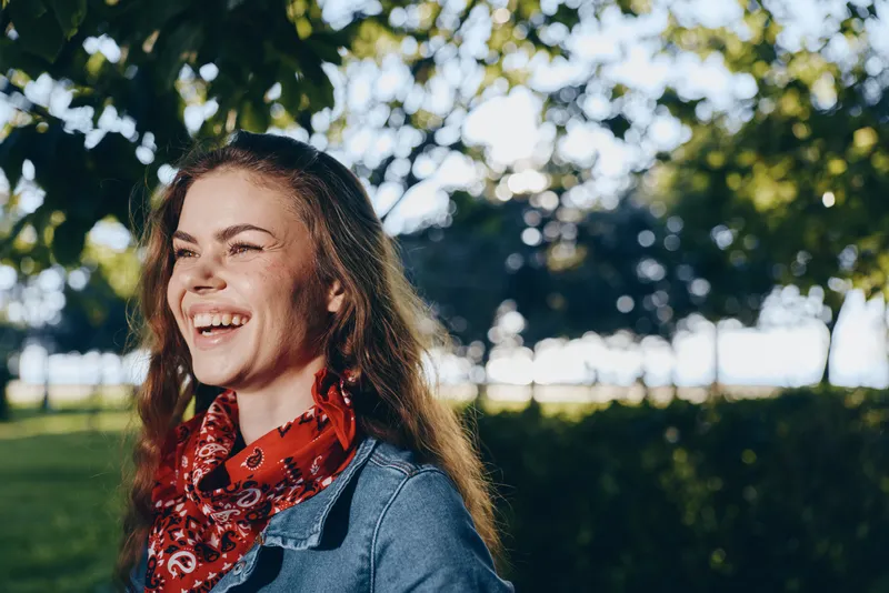 smiling woman with red bandana outdoors laughing happily, casual jacket, natural sunlight, green park background, joyful mood in warm weather