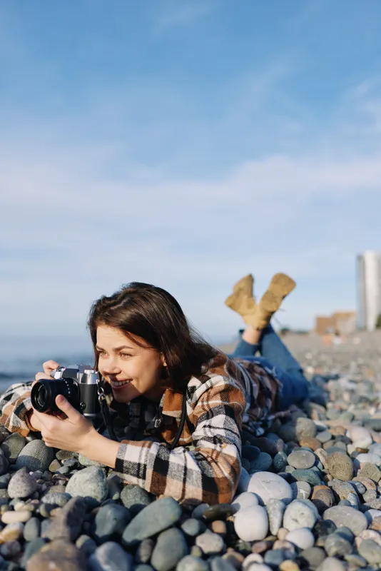 Photographer woman lying on rocky beach captures candid moments with camera among pebbles and blue sky, highlighting outdoor lifestyle, creativity and nature in a seaside scene