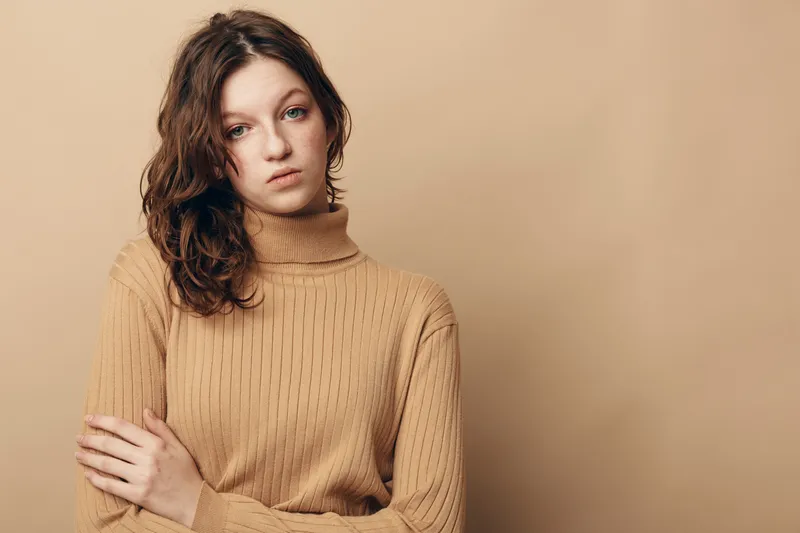 Young woman with curly hair wearing beige knitted sweater looks thoughtful and serious against plain beige background. Concept of contemplation, autumn fashion, and natural beauty.