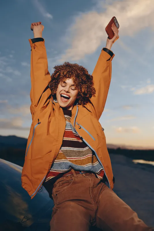 Woman outdoor lifestyle joyful sunset portrait, orange jacket and curly hair, arms raised cheering, backpack visible, warm evening sky with distant hills, energetic mood, on mountain trail