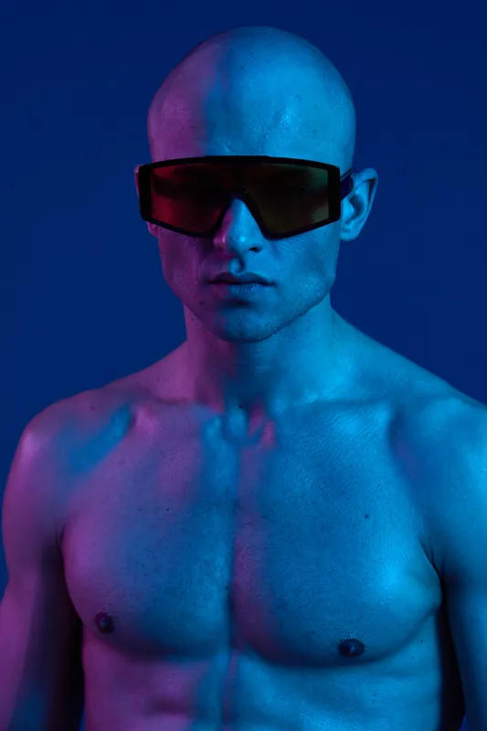 A striking man with a shaved head and sunglasses, illuminated by vibrant neon blue light, displaying a unique, artistic look. Perfect for modern fashion concepts.