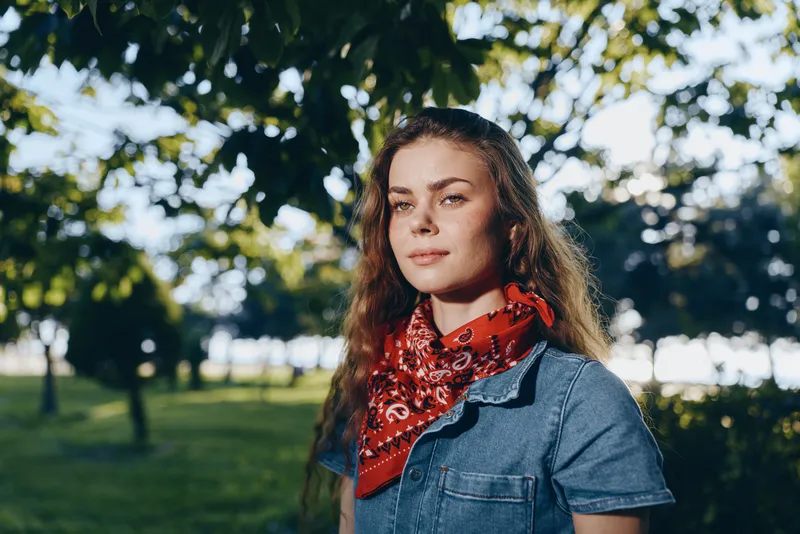 lifestyle woman in modern boho-western denim street style with red bandana stands outdoors in soft film color light, natural background, summer greenery, casual fashion