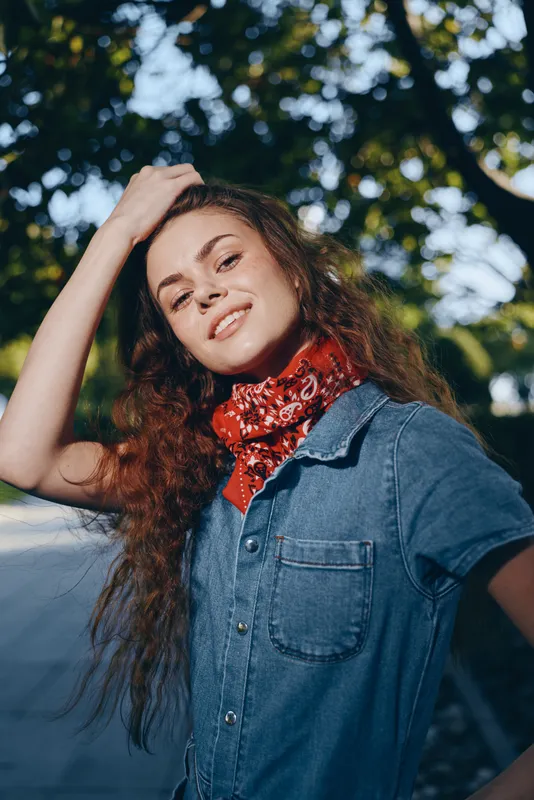 Lifestyle woman in modern boho-western denim street style with a red bandana, natural makeup, and relaxed pose, captured in warm film color under leafy sunlight.
