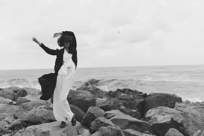 Woman on a rocky shoreline, wind blowing her coat and scarf, moving gracefully by the sea in a dramatic fashion black and white composition