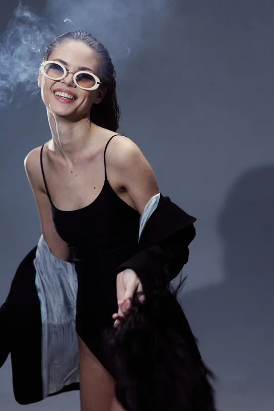 Smiling woman wearing sunglasses and black dress with jacket, enjoying a playful and confident moment in studio lighting.