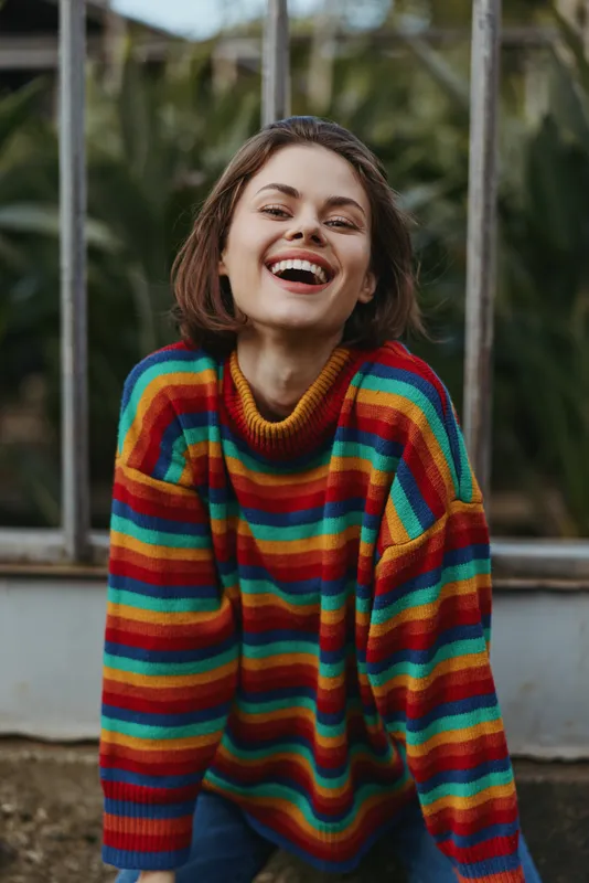 Young woman in her 20s wearing a colorful rainbow sweater and short hair, laughing and leaning forward in an outdoor greenhouse garden setting for joyful lifestyle portrait session.