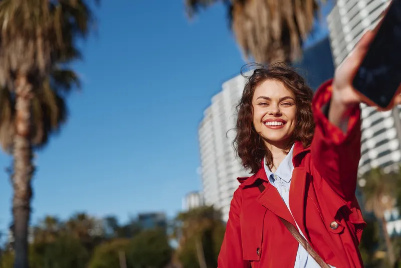 Young woman with curly hair wearing a red coat smiles brightly while taking a selfie outdoors on a sunny day with palm trees and buildings in the background.