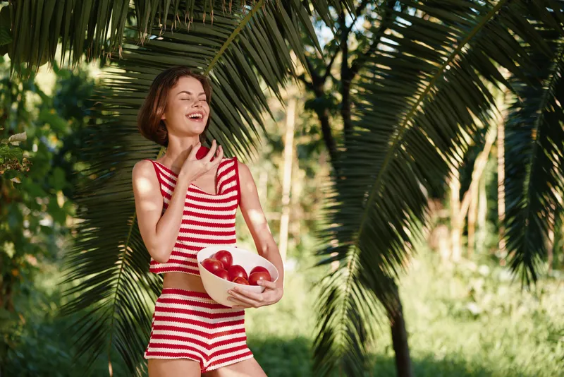 A cheerful woman in a red and white striped swimsuit walks outdoors, holding a bowl of strawberries. She smiles brightly among lush palm trees, radiating sunny summer vibes.