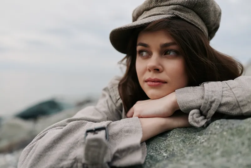 Young woman in beige coat and hat leans on stone outdoors, looking sideways with thoughtful expression. Overcast sky and blurred background create calm, contemplative mood.