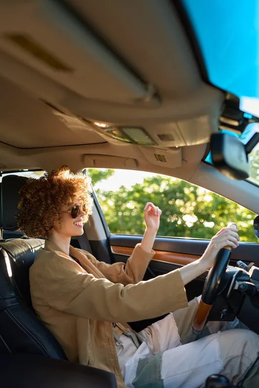 Young woman with curly hair drives a car wearing sunglasses and a beige coat, enjoying a sunny day with the bright view outside the window