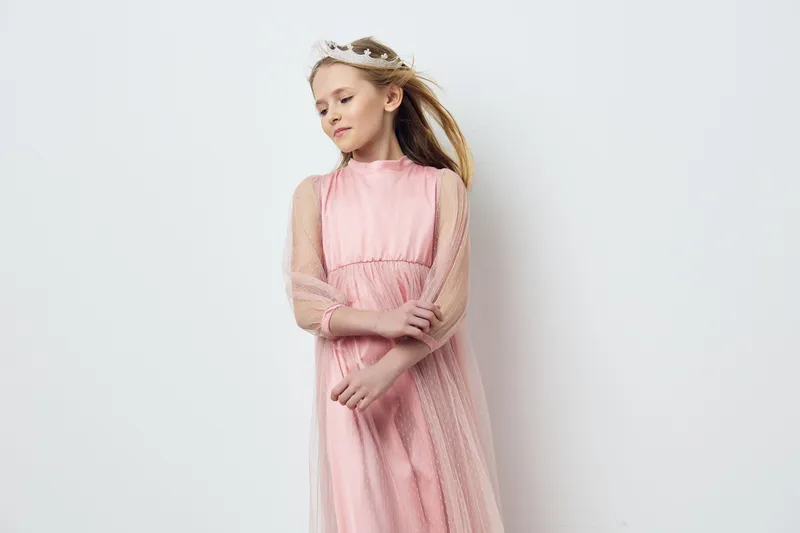 Elegant young girl in a pink dress, expressing a serene and dreamy emotion against a soft white background, ideal for fashion and childhood themes