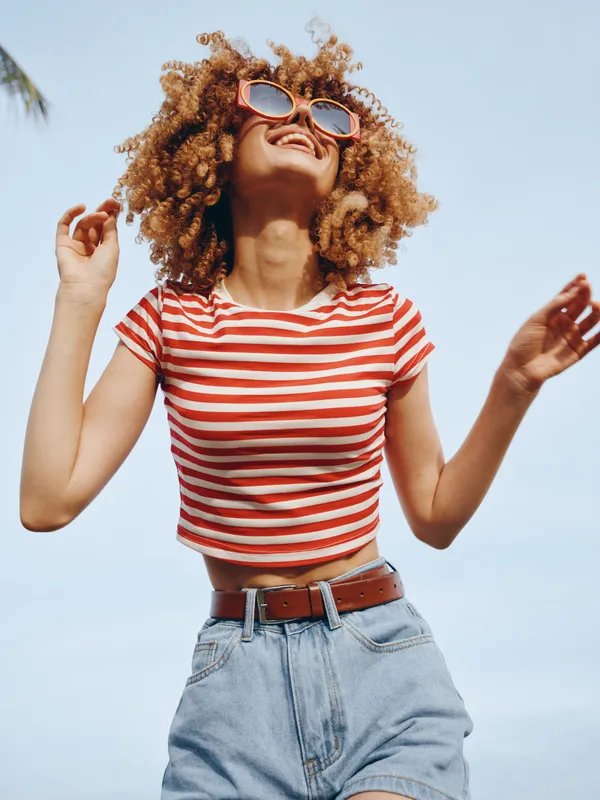 Vibrant outdoor portrait of a young woman in a red and white striped crop top, denim shorts, and sunglasses, smiling as she enjoys a sunny day.