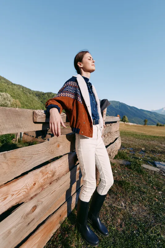 Woman standing by a wooden fence in the countryside with mountains and sunlight, wearing a cozy sweater and boots. Portrait of relaxed outdoor fashion, serene nature and clear sky moment.