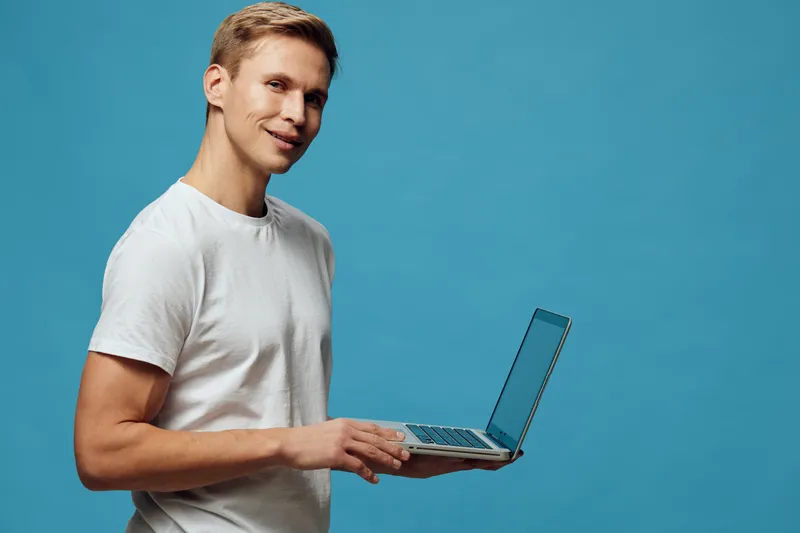 Man holding laptop computer with open screen in hand, smiling at camera, dressed casually in light t-shirt, standing against plain blue background. Studio portrait, technology and lifestyle concept.