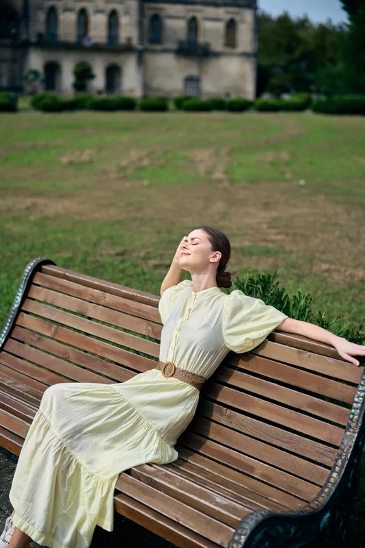 A woman in a pale yellow dress relaxes on a wooden bench in a park, eyes closed, enjoying warm sunlight and a peaceful, contemplative moment.