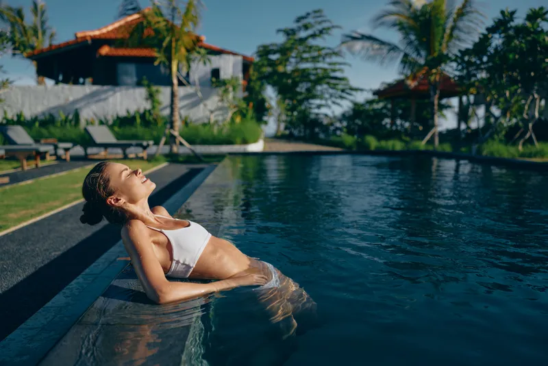 Relaxed woman lounges at pool edge in a tropical resort, soaking sunlit water, enjoying quiet vacation mood, swimsuit, serene outdoor leisure scene, blissful escape
