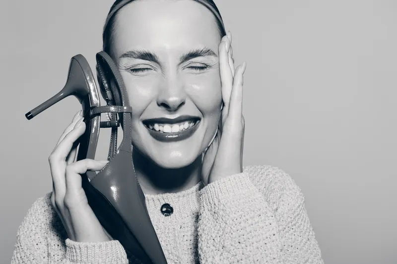 Happy young white woman with light skin holds high heel shoe near face, eyes closed and smiling in cozy sweater. Concept of fashion, joy, and female beauty with casual style.