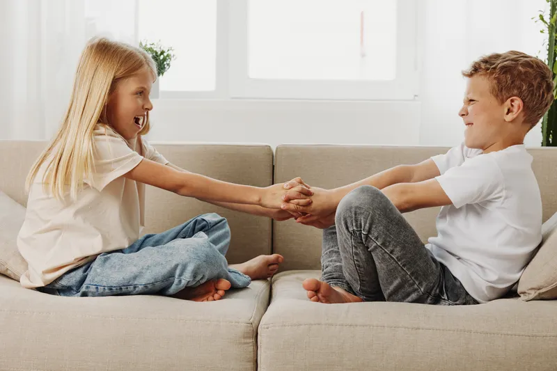 Two children sitting on beige sofa holding hands and smiling at each other in bright living room. Concept for friendship, childhood joy, family bonding and playful moments.