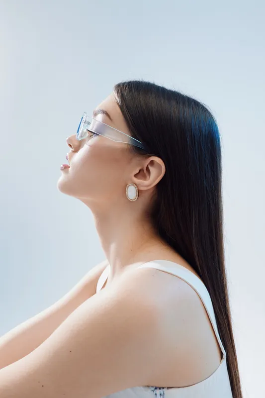Profile of a young woman with long dark hair wearing transparent glasses and white earrings. Soft lighting highlights her smooth skin and elegant beauty. Concept for fashion and beauty.