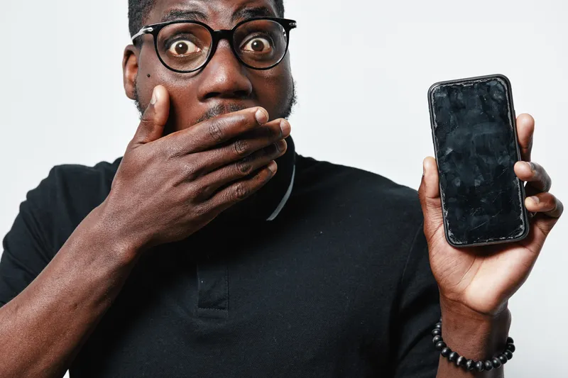 Black man with dark skin and glasses showing broken smartphone with shocked expression on white background. African ethnicity, technology damage and surprise concept.