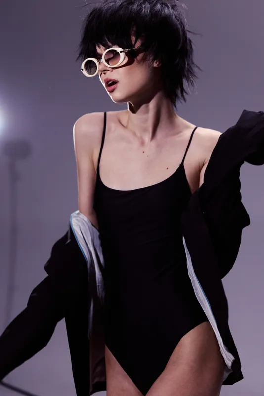 Stylish woman wearing sunglasses and black bodysuit with jacket, posing confidently in moody lighting and modern fashion style.