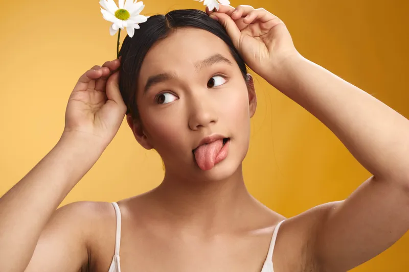 asian woman with a flower headband in a bright studio, playfully sticking her tongue and smiling, creating a lighthearted, vibrant mood and a fresh, youthful atmosphere.