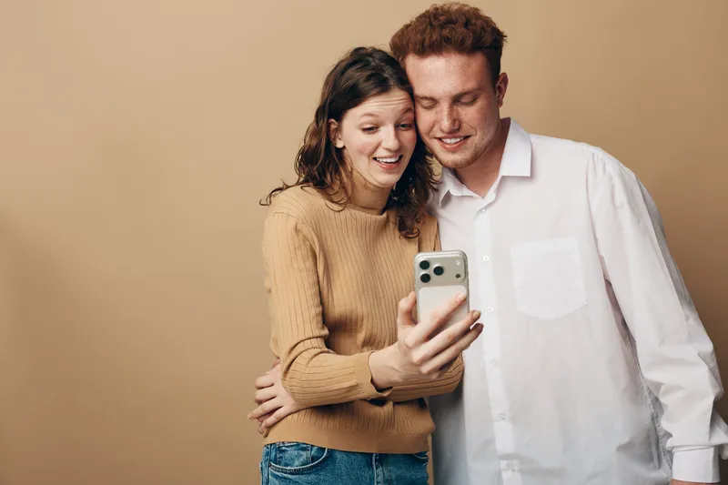 Young couple takes selfie with smartphone smiling and hugging on beige background. Casual clothing and happy mood create concept of friendship, technology, and modern lifestyle.