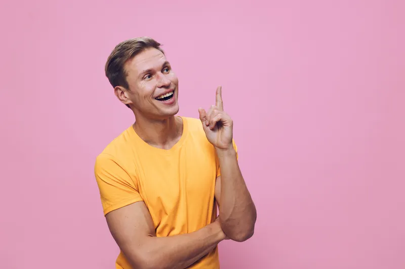 Smiling young man in bright yellow t-shirt pointing upwards with happy expression on pink background. Studio portrait of cheerful male person with positive emotions and casual style. People lifestyle 