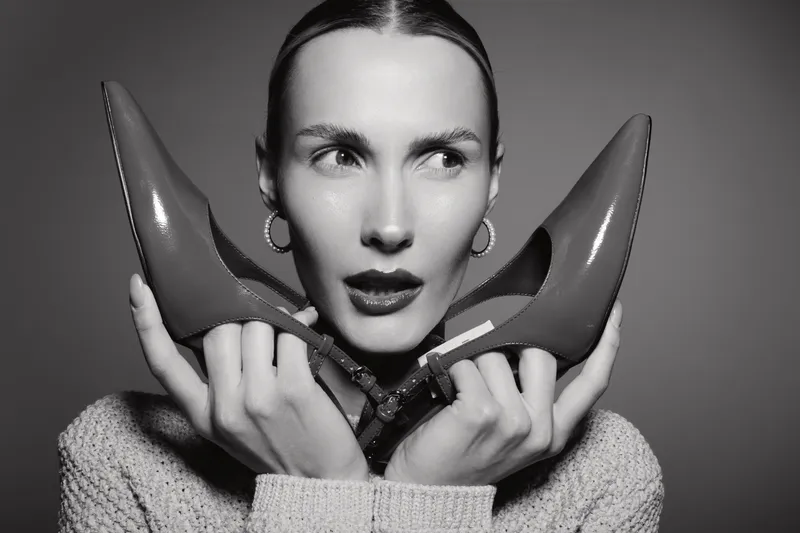 Caucasian woman with light skin holds high heel shoes near face, wearing hoop earrings and knitted sweater. Concept of fashion, style, and footwear.