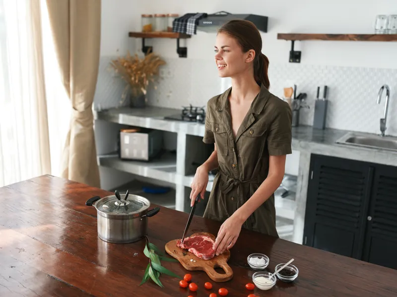 Женщина на кухне улыбается, готовит ужин из мяса и овощей на деревянном столе на фоне стильной кухни