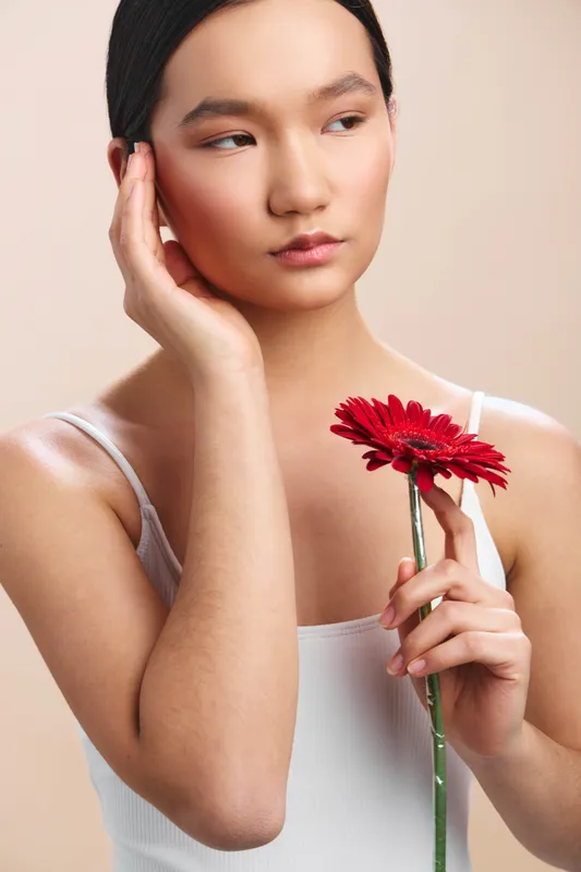 asian woman in soft studio light holds a red flower near her face, thoughtful expression, conveying calm elegance and contemporary beauty in a minimalist setting.