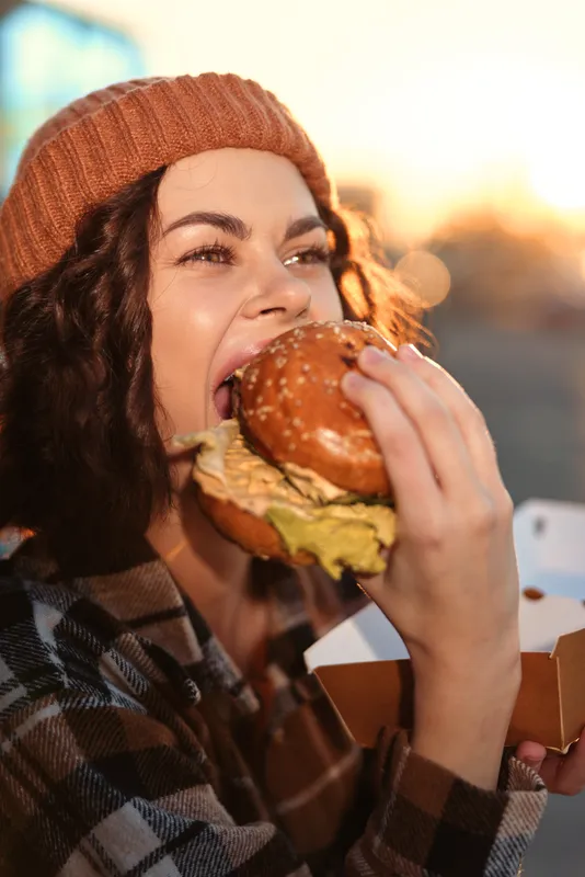 Woman eating burger outdoors, smiling while holding a takeaway box at golden hour. Candid lifestyle portrait with authenticity, mindful living and emotional storytelling in urban warmth.
