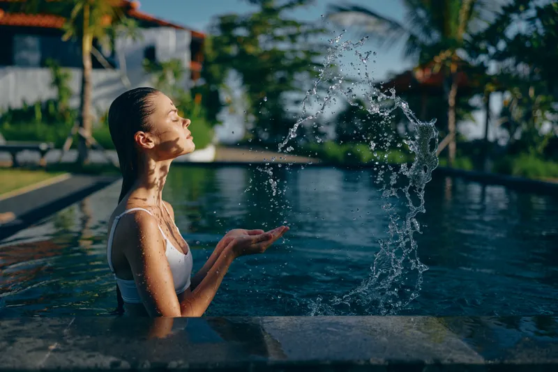 Young woman in white swimsuit playing with water near outdoor pool. Relaxed lifestyle scene with tropical background and natural sunlight creating a fresh mood.