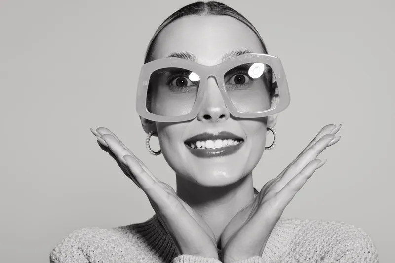 Smiling Caucasian woman with pale skin wears oversized glasses and knitted sweater posing with hands near face on grey background. Concept of fun and fashion.