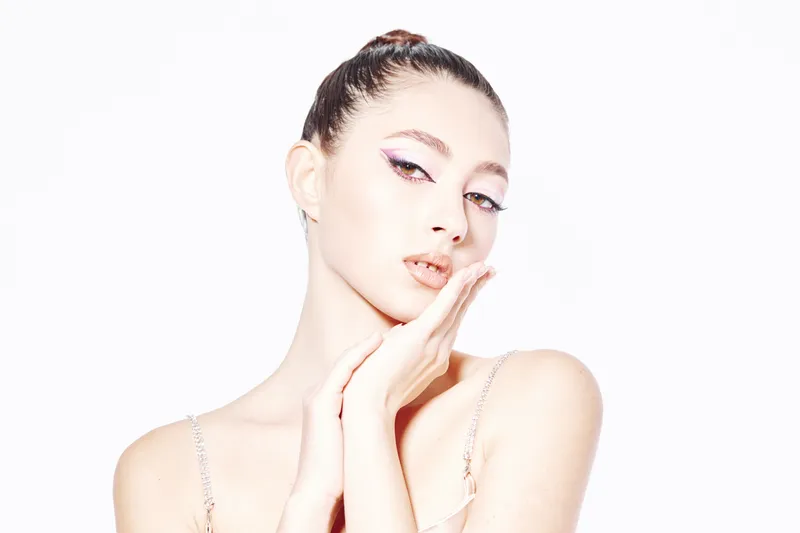 Young woman with creative colorful eye makeup poses with elegant hand near face on white background. Fashion beauty concept expressing modern feminine style and clean skin care.