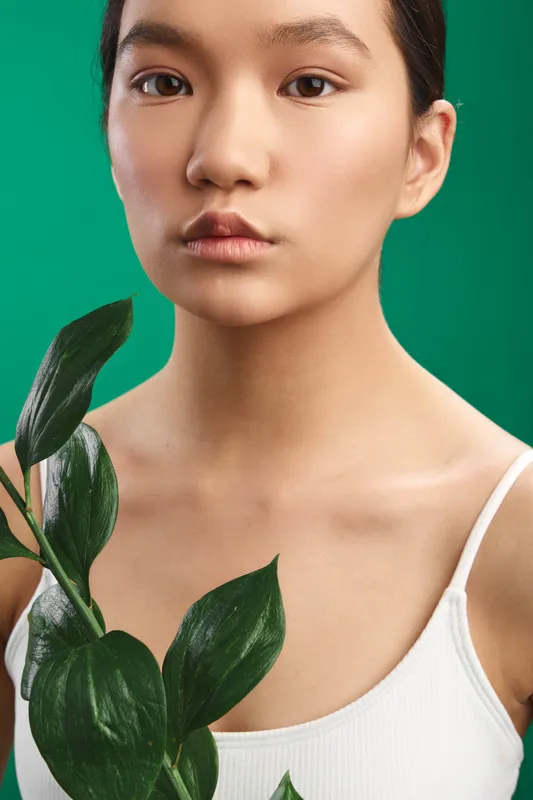 asian woman gazes softly at the camera while holding leafy greens in a bright studio, soft lighting and neutral tones emphasize simplicity, natural beauty, and a calm, fresh mood.