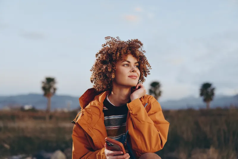 Woman lifestyle outdoors, curly hair, jacket, smartphone, field, sunset, portrait, orange coat, smiling softly, relaxed stance, gentle breeze, rural landscape, blue sky