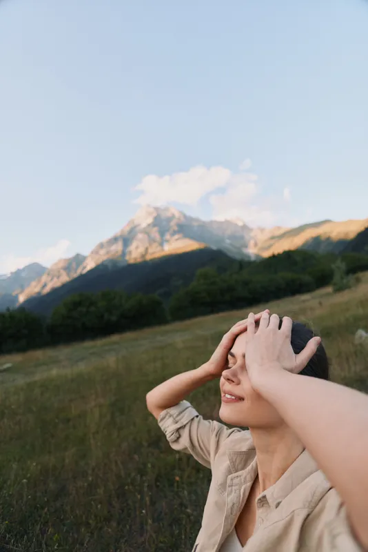 Young woman outdoors in nature, smiling with hands on head, enjoying mountain landscape, clear sky, casual clothing, peaceful moment, natural light