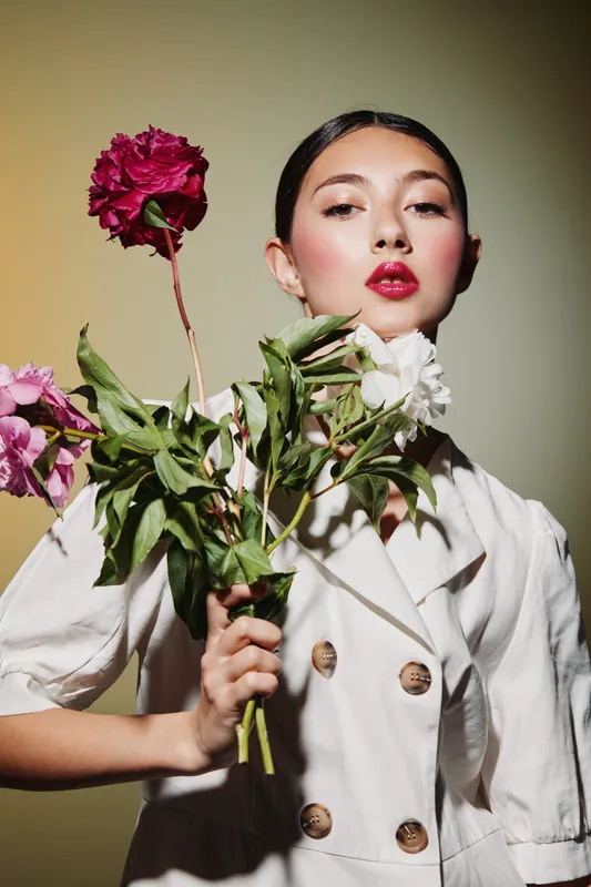 Young woman with glossy makeup and bold red lips holds colorful flowers against a soft yellow background. She wears a buttoned white blazer. Concept of beauty fashion creative portrait.