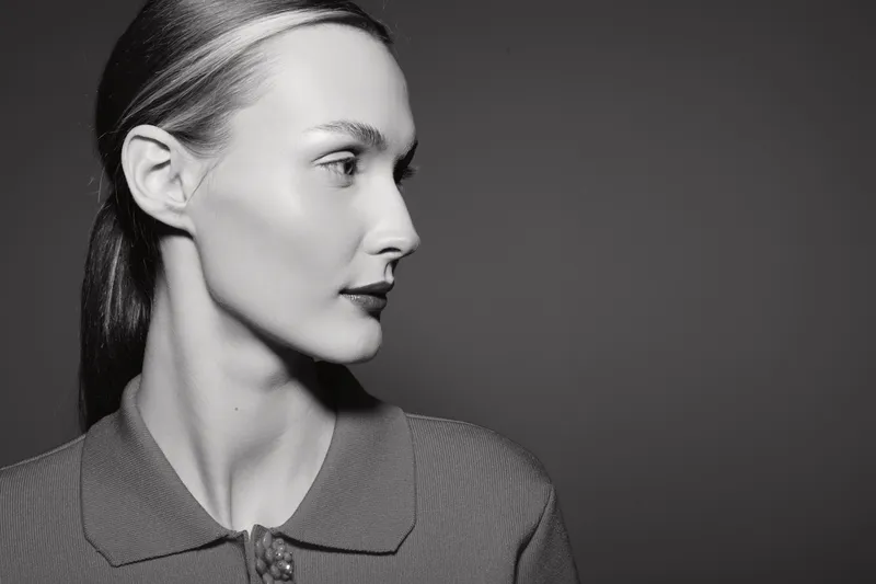 Profile portrait of a young white woman with fair skin and slicked back hair wearing a textured shirt on a dark background.