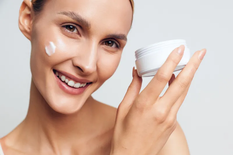 Smiling woman with clear skin holding white cream jar and applying face moisturizer on cheek. Beauty skincare concept with natural look and soft lighting on white background.