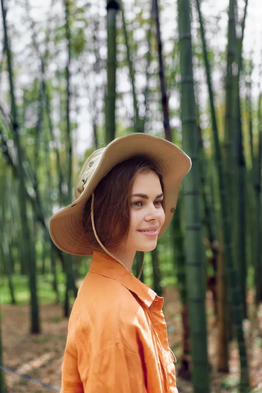 Woman portrait with hat and smile in bamboo forest, outdoor lifestyle image of young female in orange shirt, relaxed pose with natural light and shallow depth of field.