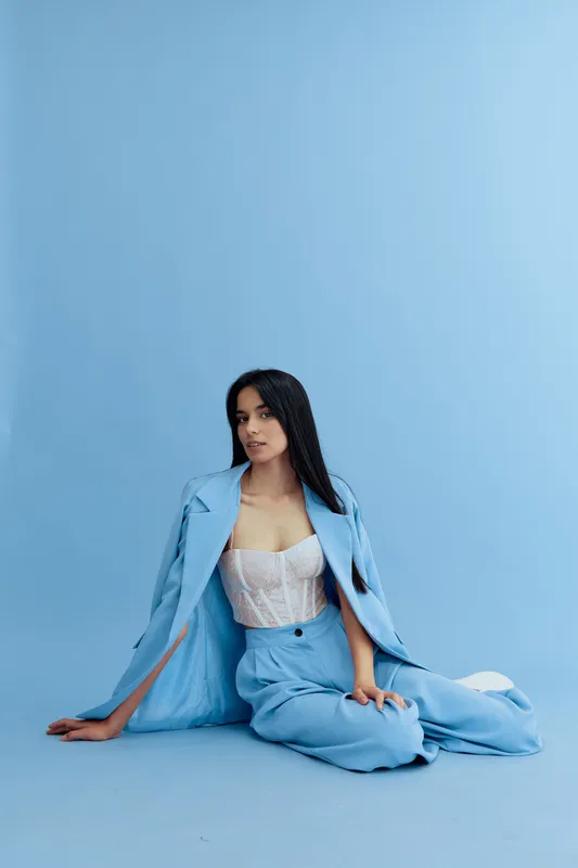 Young woman in light blue suit sitting on floor against pastel blue background, wearing white lace corset, with long black hair and relaxed calm expression.