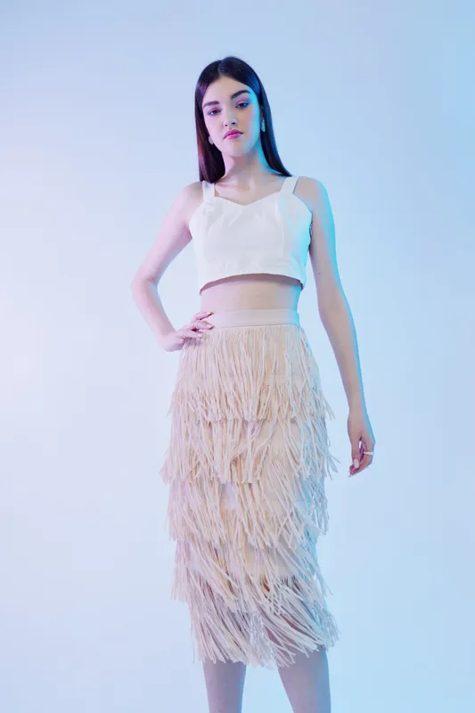 Young woman beauty fashion model posing in a white crop top and beige fringed skirt. Studio shot with soft lighting on a gradient blue background. Concept for style, elegance, and modern trends.