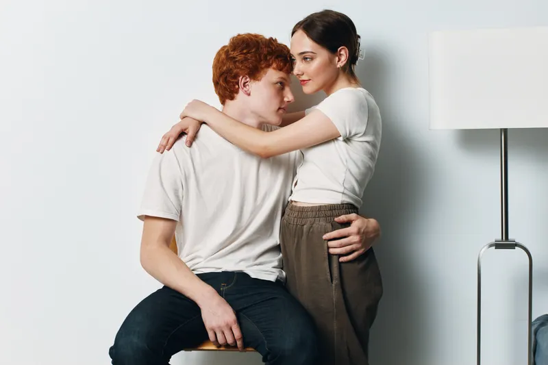 Couple, love and intimate moment with young woman embracing man sitting on chair for affection and relationship in home interior. Romance, tenderness and casual clothing with lamp on white wall.