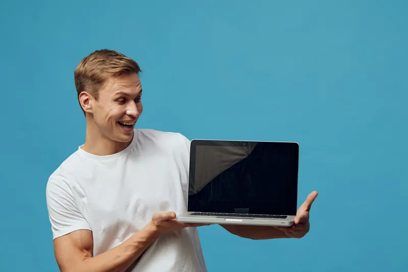 Smiling young man holding open laptop computer with blank screen isolated on blue background in studio. People lifestyle concept.