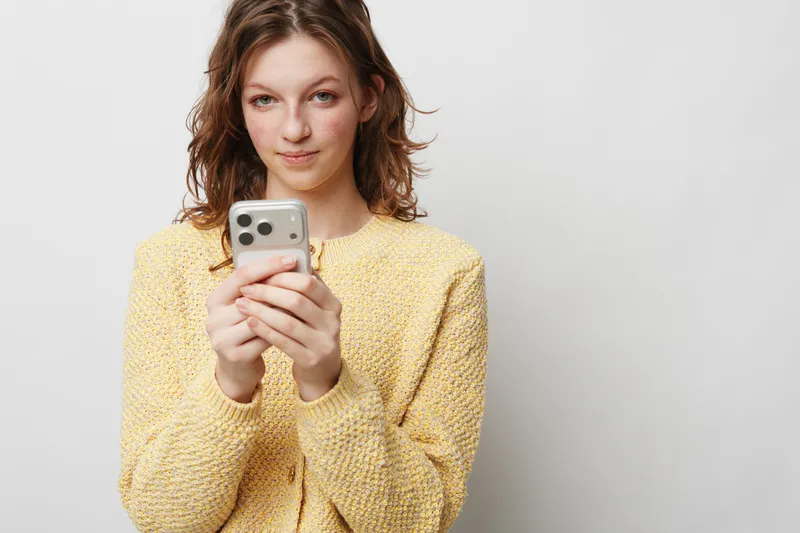Young woman in yellow sweater holds smartphone and looks at camera with slight smile. Isolated on white background. Concept of communication, technology, and casual lifestyle.